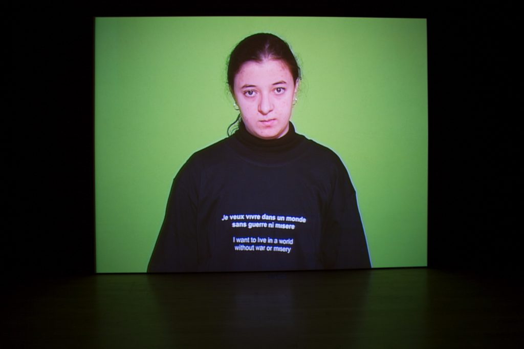 An image of a woman in front of a green background wearing a sweatshirt that says "I want to live in a world without war or misery" in both English and French