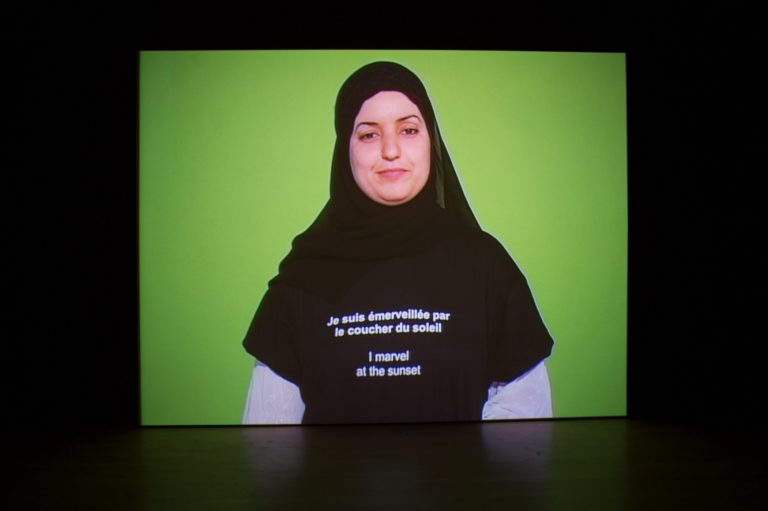 An image of a woman standing in front of a green background wearing a hijab and a sweatshirt that says "I marvel at the sunset" in both English and French.