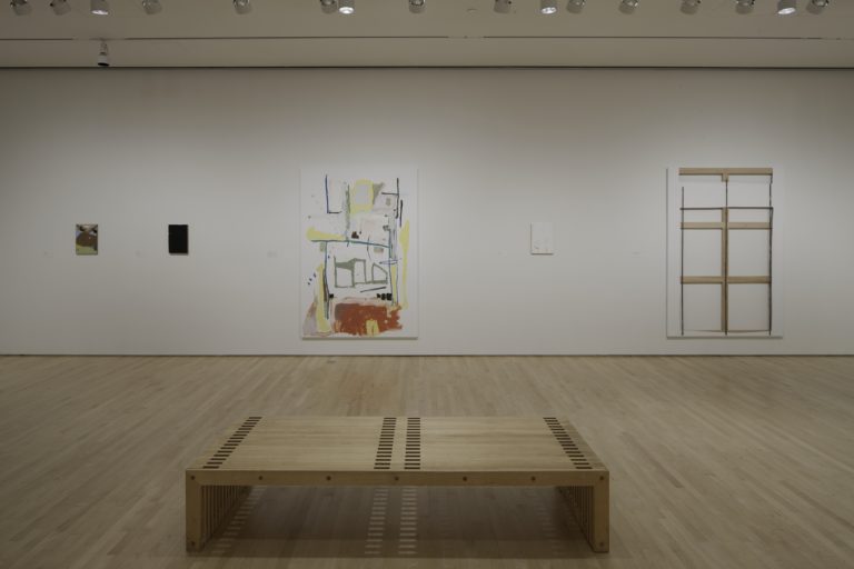 3.)	A white room with four pieces of art work by Richard Aldrich on the walls. There is a bench in the middle of the room.