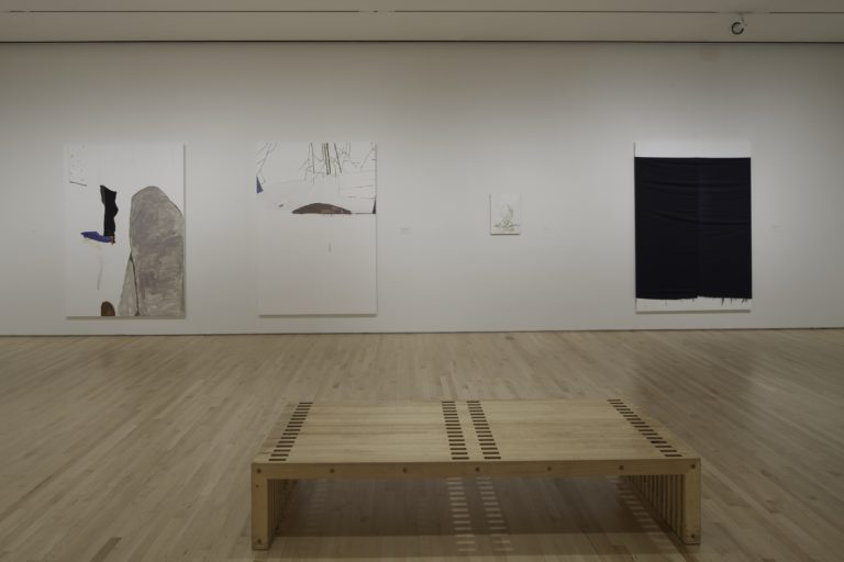 A white room with four pieces of art work by Richard Aldrich on the walls. There is a bench in the middle of the room.