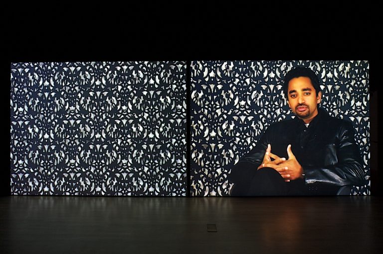 An image of a man sitting in front of a patterned wall