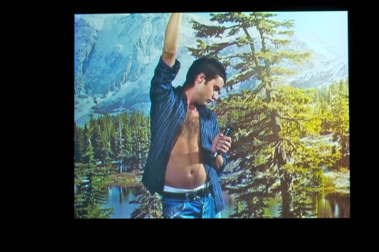 A man with one arm raised above his head and the other holding a mic, wearing an open button down that exposes his chest, standing in front of a backdrop showing a landscape with moutains and trees.