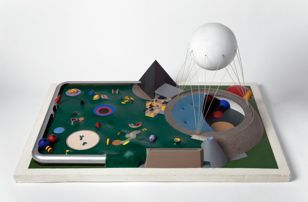 Table top model of a brightly colored pavilion with a large sphere suspended above, set into a lawn