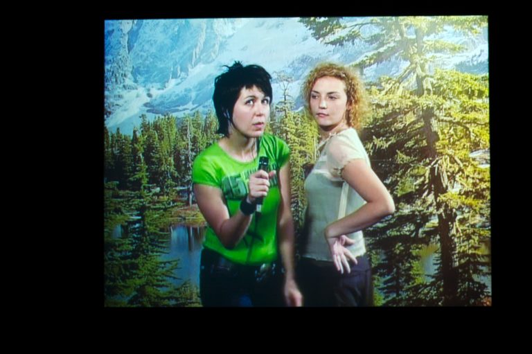 Two women, one holding a mic, standing in front of a landscape backdrop