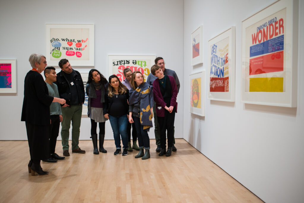 A group of visitors lean in to get a better look at silkscreened prints by Corita Kent