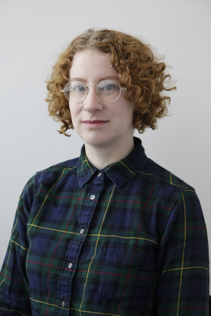 A woman with curly red hair and wire-rimmed glasses sits looking at the camera