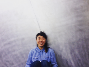An Asian-American woman with bangs and wearing a denim shirt sits before a gradated grey background, Geraldine Ah-Sue