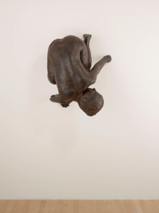 A bronze sculpture of a crotching figure with glass eyes hung upside down from a white wall