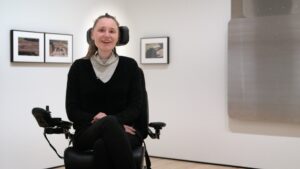 Artist Park McArther sitting in her wheelchair, with photographs on the wall behind her