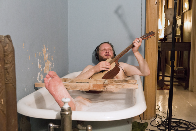 A naked Caucasian man plays the guitar in a bathtub, Kjartansson