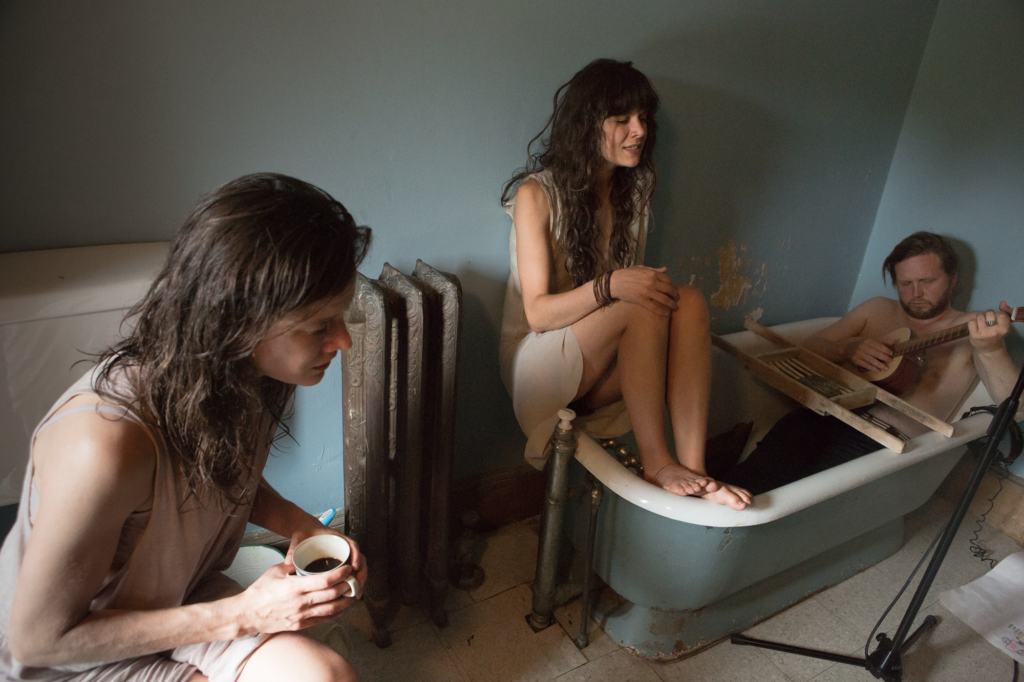 Two Caucasian women and a bearded man sit around a bathtub; the man holds a guitar, Kjartansson