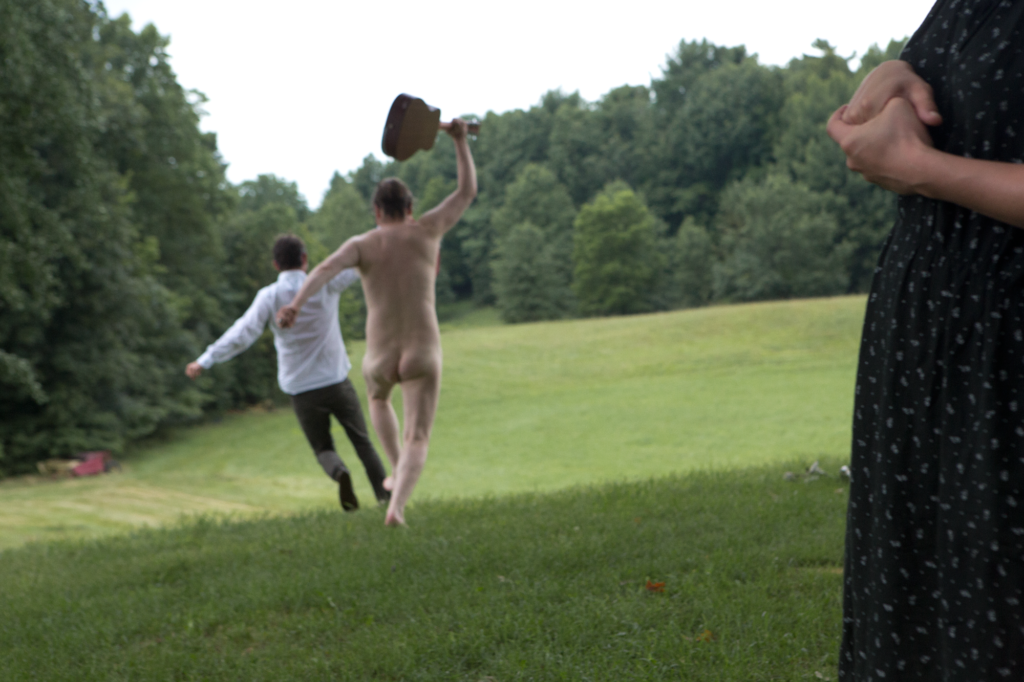 A naked Caucasian man holding a guitar chases another clothed man through a green field, Kjartansson