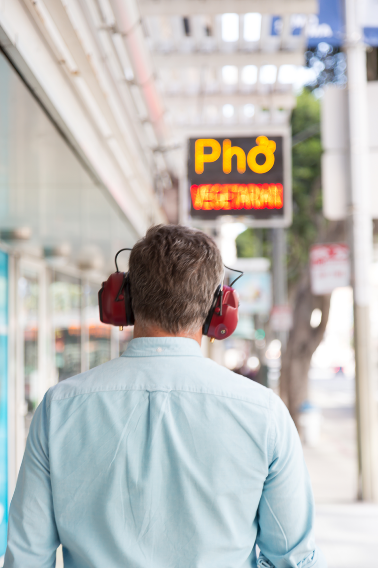 A man wearing red headphones is seen from behind walking on a street toward a sign saying "Pho", Kubisch Soundtracks