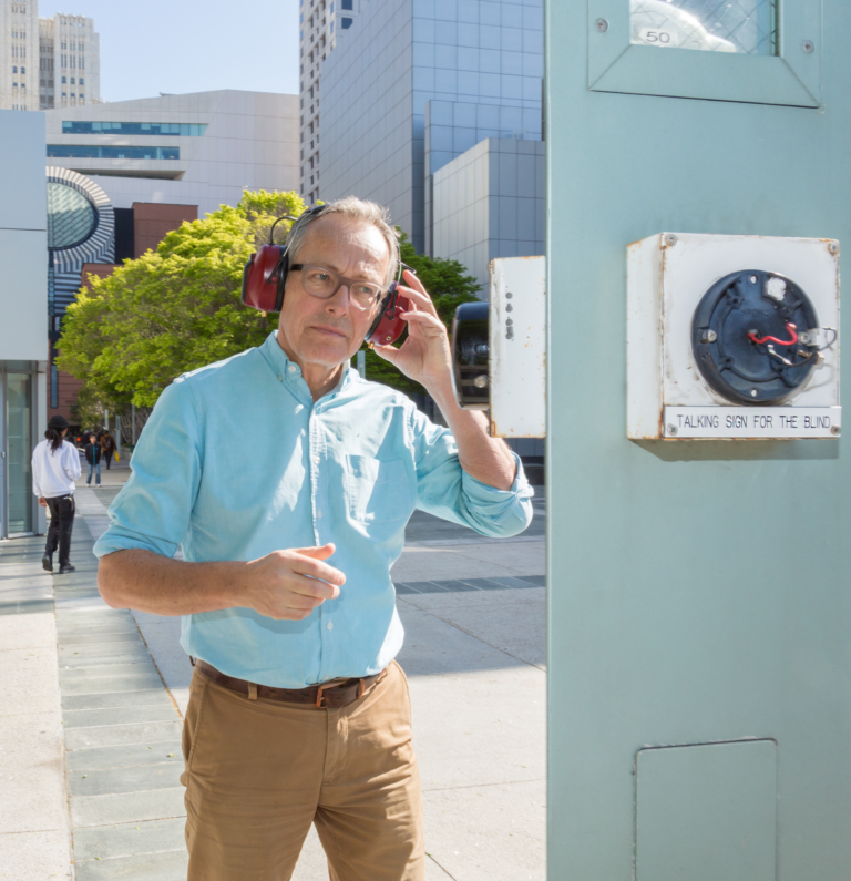 A Caucasian man wearing red headphones and glasses walks toward an electrical meter on the street, Kubisch Soundtracks