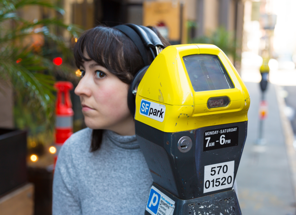 A Caucasian woman with brown hair appears to be listening to a parking meter, Kubisch Soundtracks