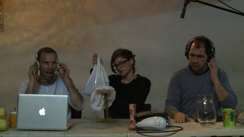 Two Caucasian men and one women sit at a table with a computer and various other items, Ben Ner Soundtracks