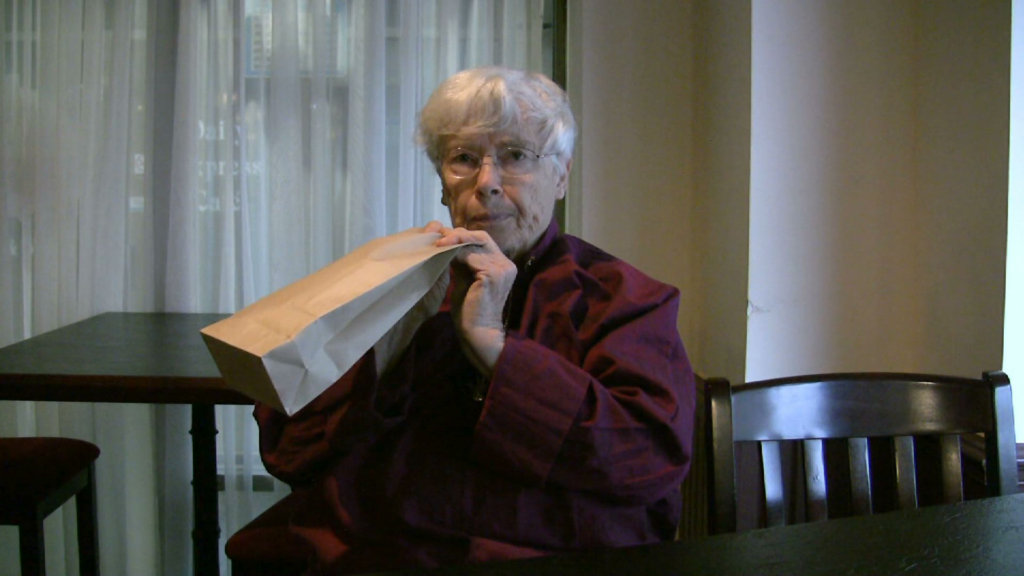 An elderly Caucasian woman with white hair holds a paper bag to her face, Lozano-Hemmer Soundtracks