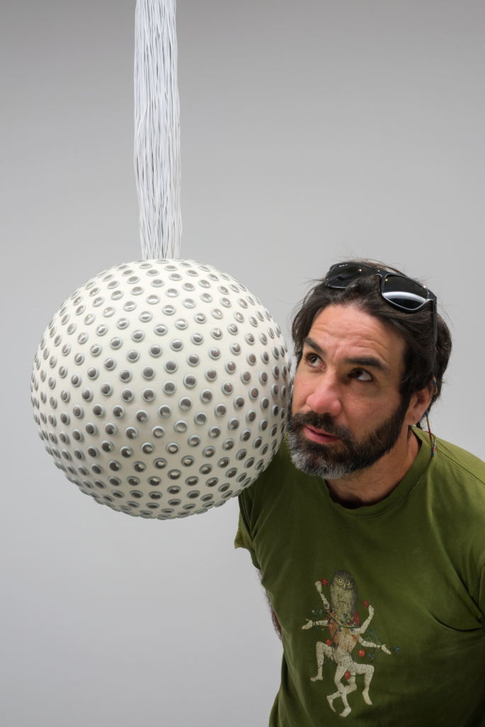 A Caucasian man with dark hair and beard listens to a perforated hanging sphere, Lozano-Hemmer Soundtracks