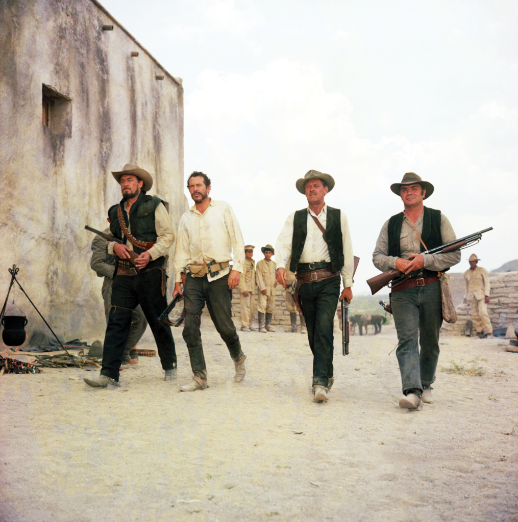 Four cowboys walking with guns through a village