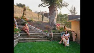 A photograph of a backyard, with a shirtless man reclining on a staircase, and a woman in high heels and a slit-leg skirt sitting on a patio chair.
