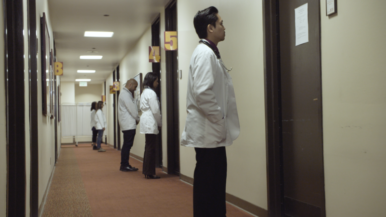 A row of doctors in white coats stand outside doors along a corridor