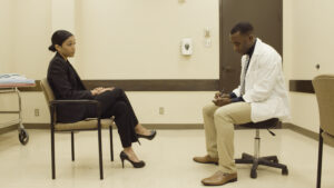 A woman in a black suit and a man in a white doctor's coat sit facing each other in a hosptial room