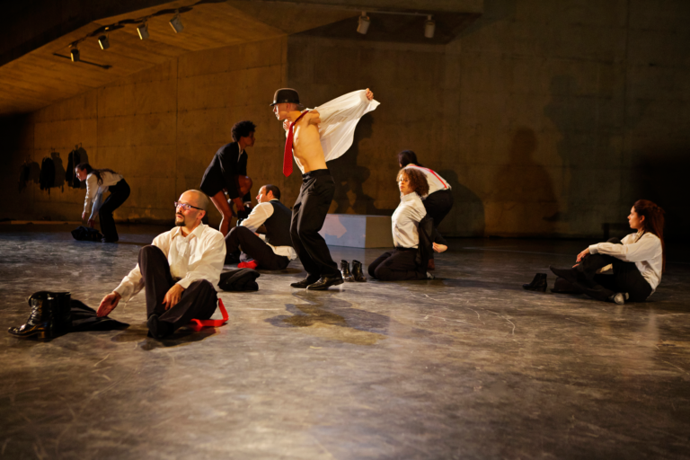 Figures wearing white shirts, red ties, and hats perform on a flat stage, Anna Halpern Soundtracks