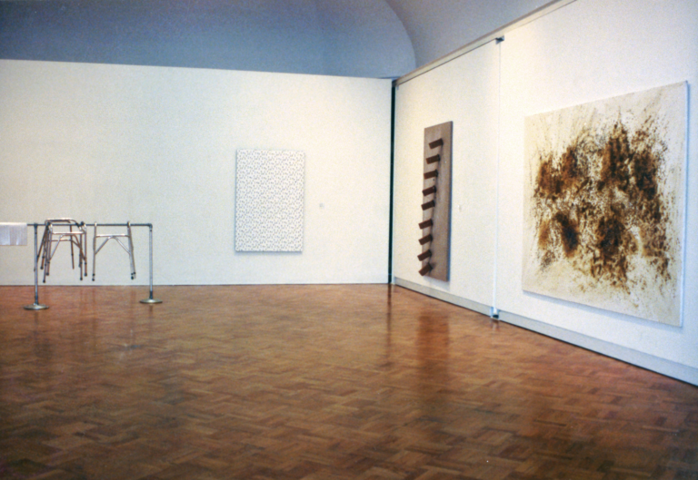 A gallery with one painting featuring dark brown smudges, a hanging steel sculpture with protruding cylinders, a hanging white artwork with a black pattern on it, and steel bars with steel furniture hanging on them. 