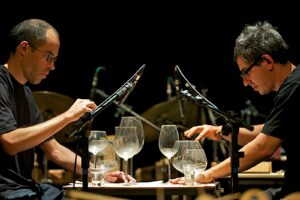 Two men play a modified glass harmonica made of wine glasses with directional microscopes