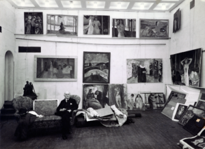 An archival black and white photograph of an elderly Caucasian man sitting in a studio surrounded by paintings