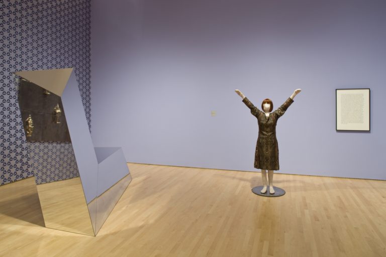 A sculpture of a woman with her hands raise din the air next to a reflective geometric sculpture and a framed block of text