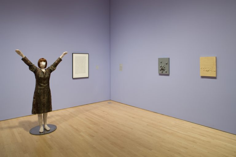 A sculpture of a woman with her hands raised in the air next to a framed block of text and two wall-hanging sculptures