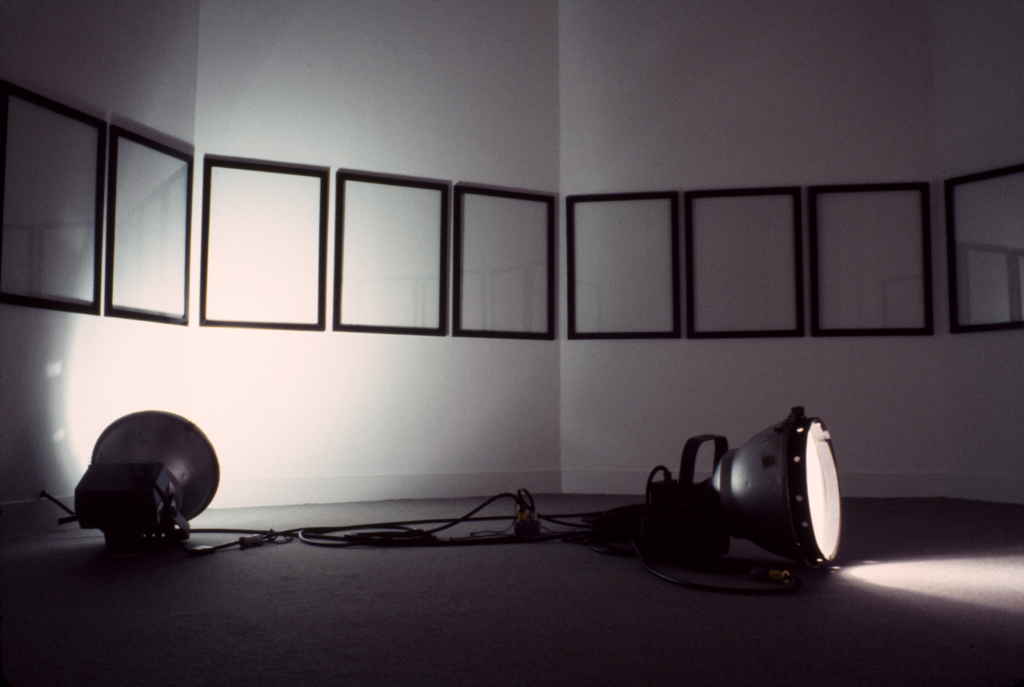 Two lamps in the foreground with a row of white canvases with black frames in the background