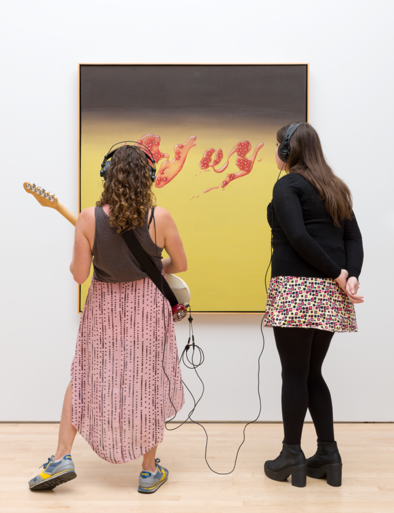 Two women, one playing a guitar and one wearing headphones, look at a painting, Kallmyer Allen, Soundtracks