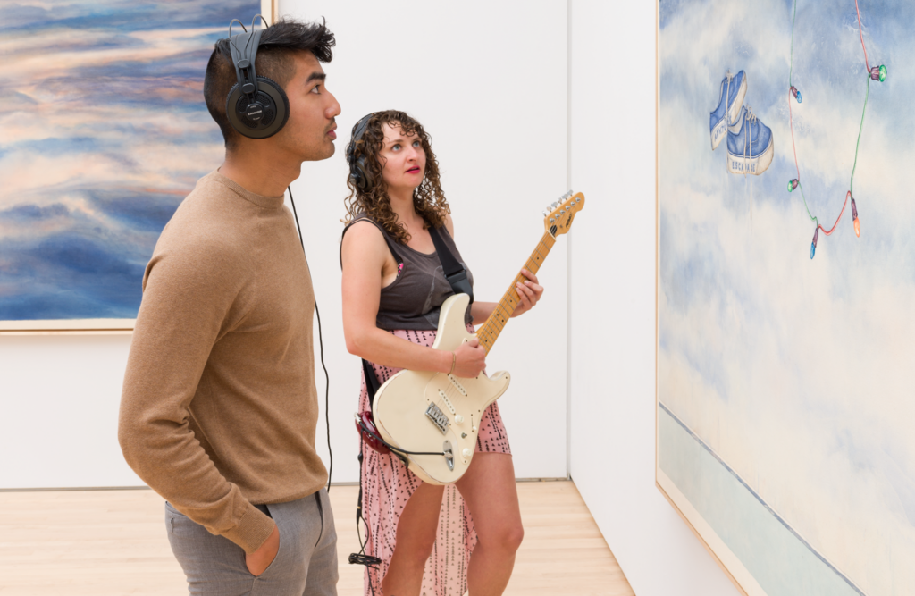 A Caucasian woman playing a guitar stand with an Asian man in headphones (listening to the guitar) looking at a painting, Kallmyer Allen Soundtracks