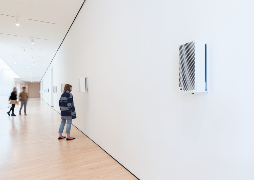 A Caucasian woman leans into a white speaker mounted on the wall, Philipsz, Soundtracks