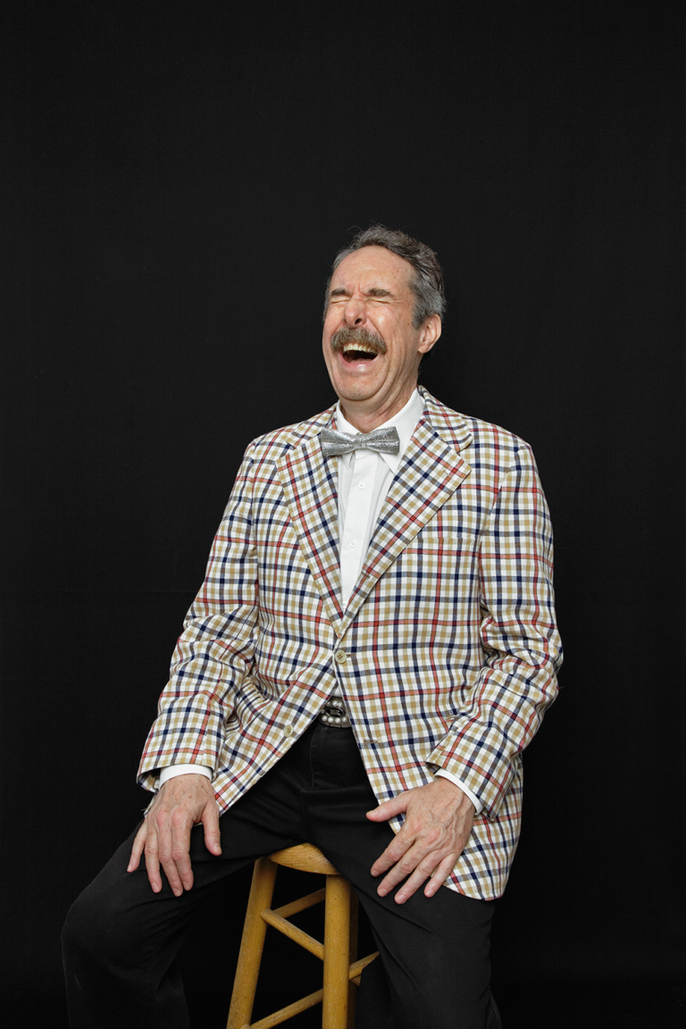 A man with salt and pepper hair, a mustache, silver bowtie and plaid jacket sits on a stool and laughs with his eyes closed