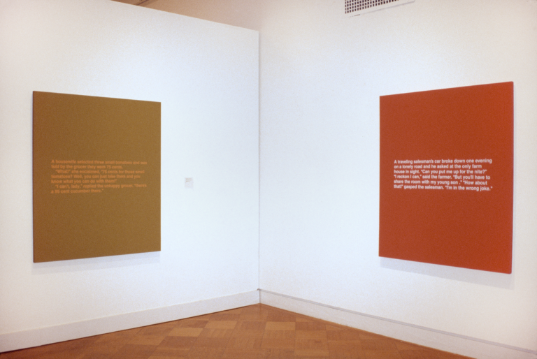 A mustard canvas and an orange canvas hanging in a gallery, each featuring blocks of white text