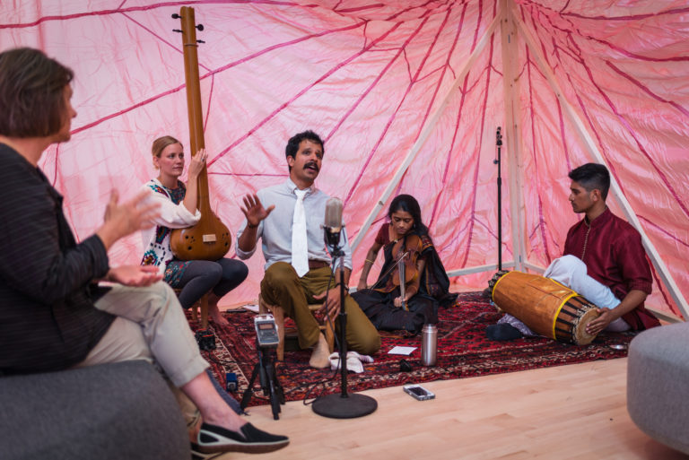 Musicians seated on a carpet with a pink fabric backdrop playing sitar, percussion and singing