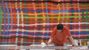 Artist Alicia McCarthy painting, with one of her giant works of painted woven lines behind her