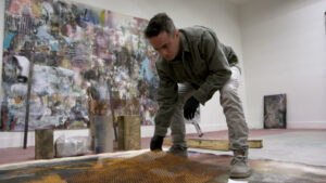 Artist Lian Everett crouching over his floor-based painting with a sheet of metal in his hand