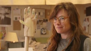 Artist Lindsey White smiling next to a glove on a stick with smiley faces drawn on the fingers