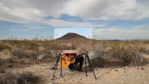 A guitarist crouching in a desert landscape behind a photograph on two tripods of that same landscape
