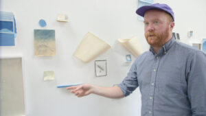 Artist Sean McFarland pointing to blue paper artworks on the wall