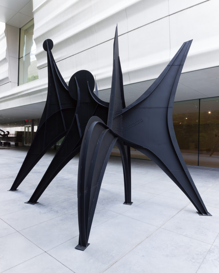 A large, standing, black metal sculpture on a terrace with the facade of SFMOMA behind