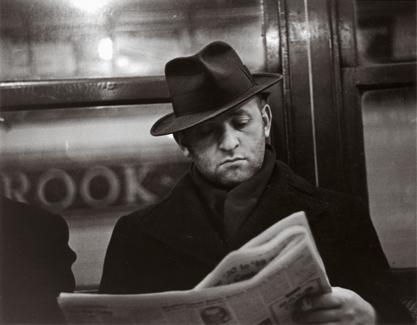 A black and white photography of a Caucasian man wearing a fedora reading a newspaper