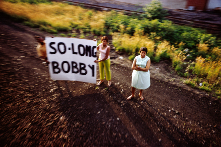 Three blurry figures look up toward the camer, two hold a banner that reads, "So-long Bobby"