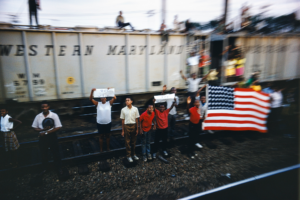 A group of people standing along a train track holding signs that say, "RFK We Love You" and and American flag