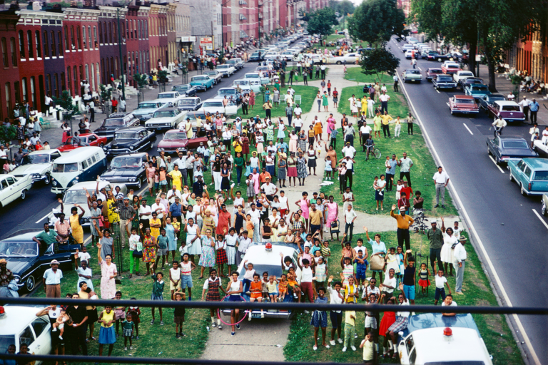 A large group of people standing on a grassy median in a city waving toward the camera