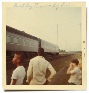 A faded square photograph of a group of black and white children and adolescents, seen from behind and in profile, watching a large train pass by from the train tracks